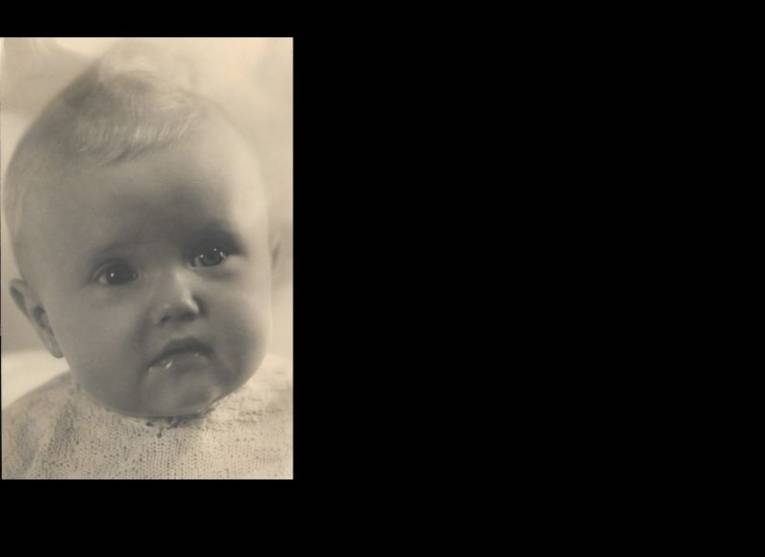 Schwarz/Weiß-Foto. Ein kleines Baby mit blondem Haar schaut in die Kamera.